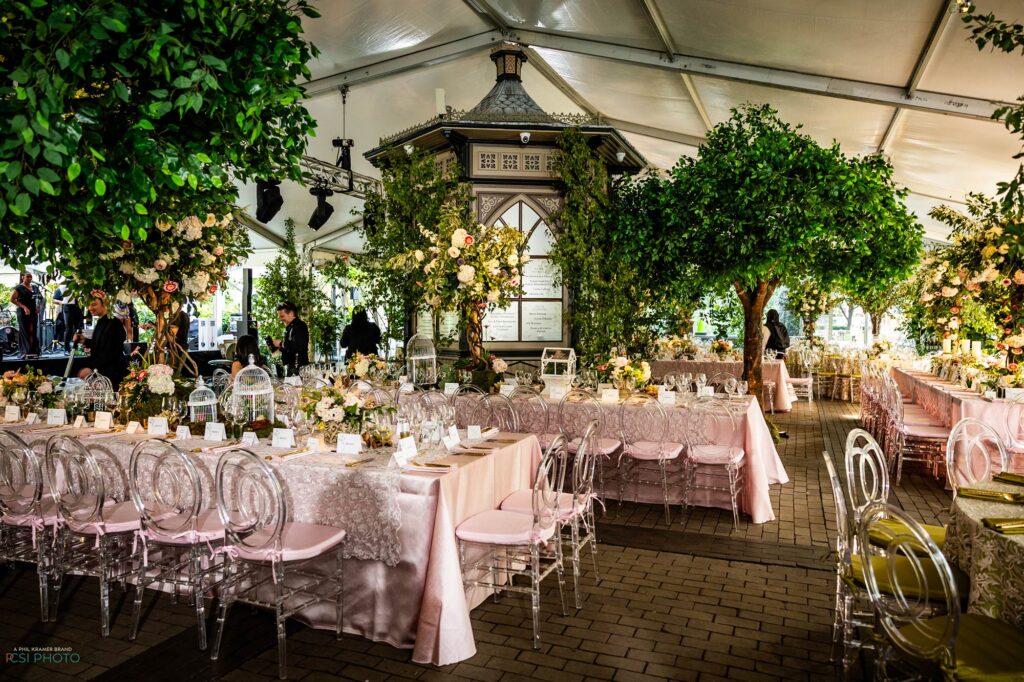 Outdoor dining event under white tent with pink linens tables, clear chairs, greenery and floral arrangements.