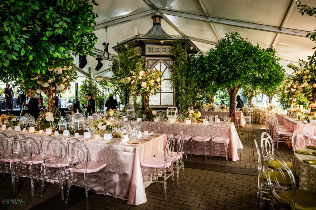 Outdoor dining event under white tent with pink linens tables, clear chairs, greenery and floral arrangements.
